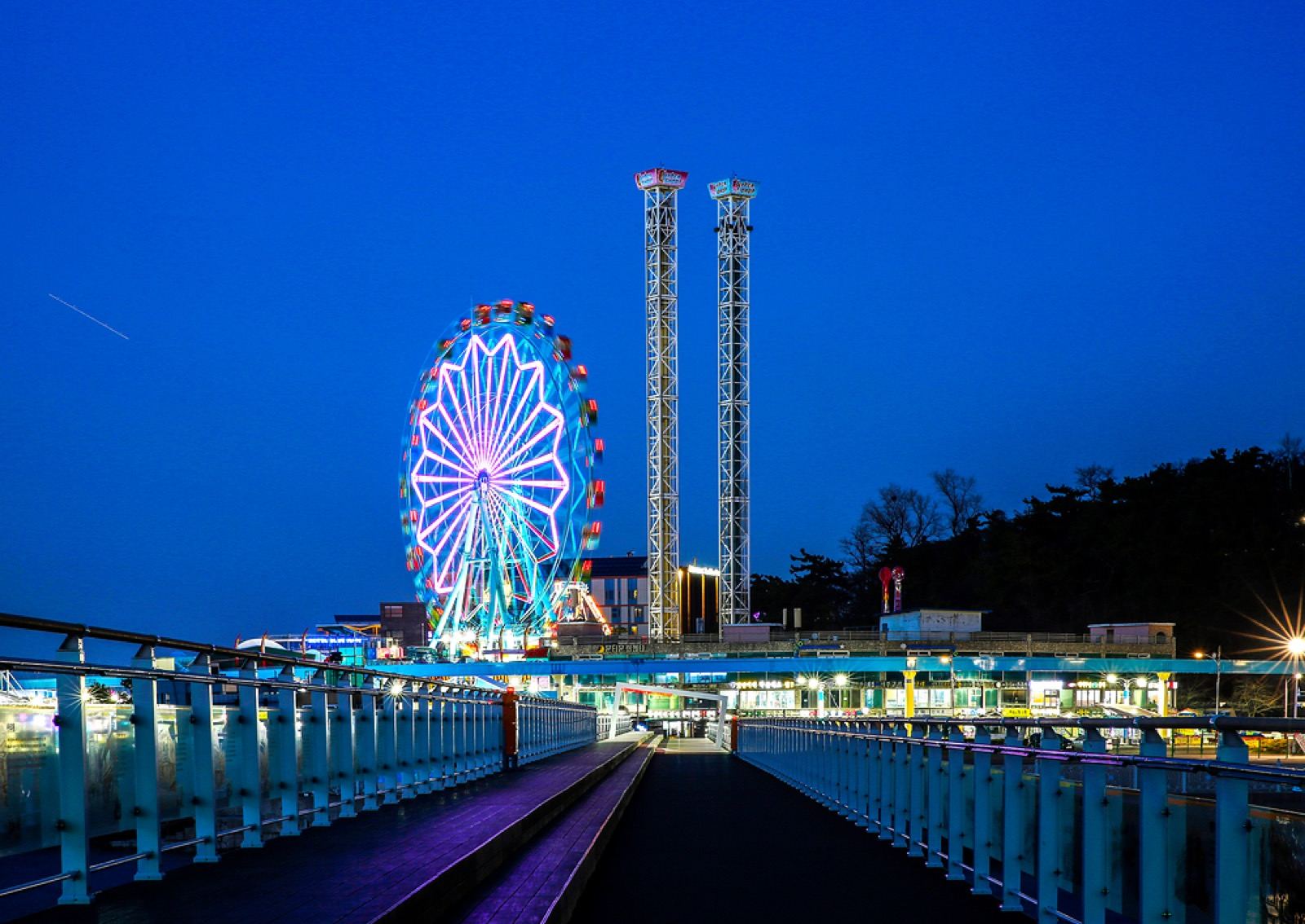 The lit Ferris wheel and drop-tower rides of Wolmido amusement park at dusk, seen from the seaside boardwalk