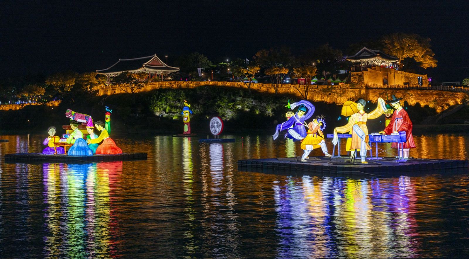Glowing figural lanterns floating on the Nam River below the lit walls of Jinju Fortress at night