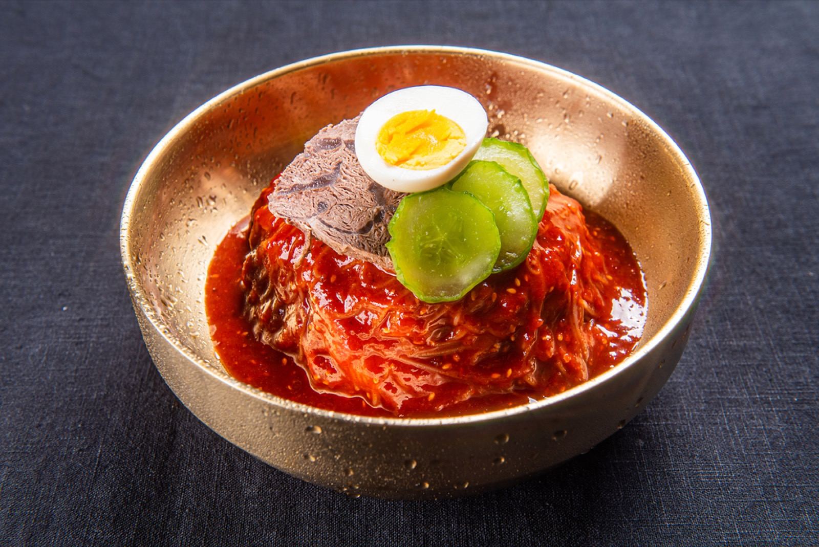 A brass bowl of bibim-naengmyeon, buckwheat noodles in bright red spicy sauce topped with egg and cucumber