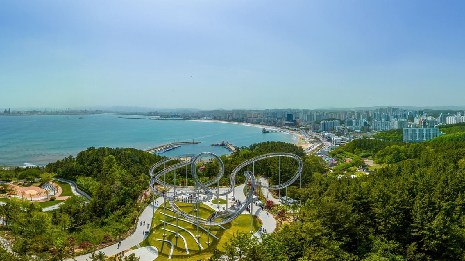 A large white steel roller-coaster-shaped walkway curving over a hilltop above Pohang