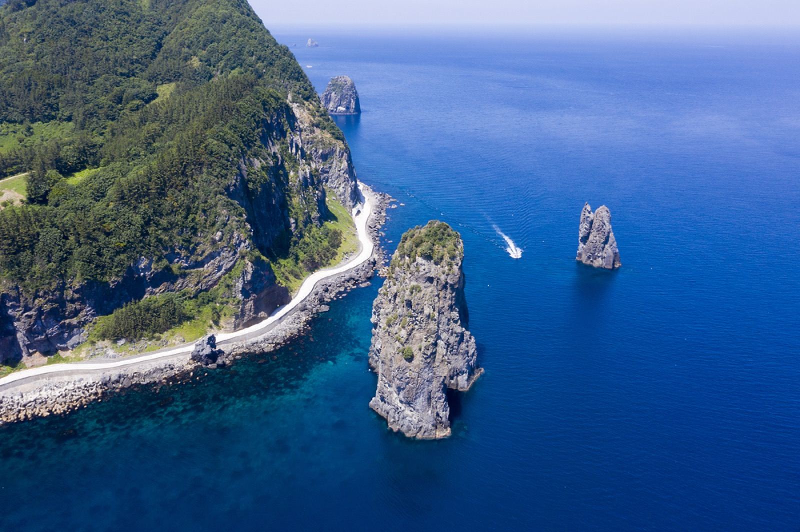 A white coastal road winding along the base of Ulleungdo's cliffs past sea stacks in blue water